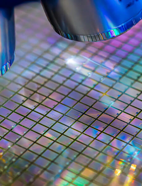 Close-up of a silicon wafer showing a grid of tiny square dies under bright, colorful lighting.